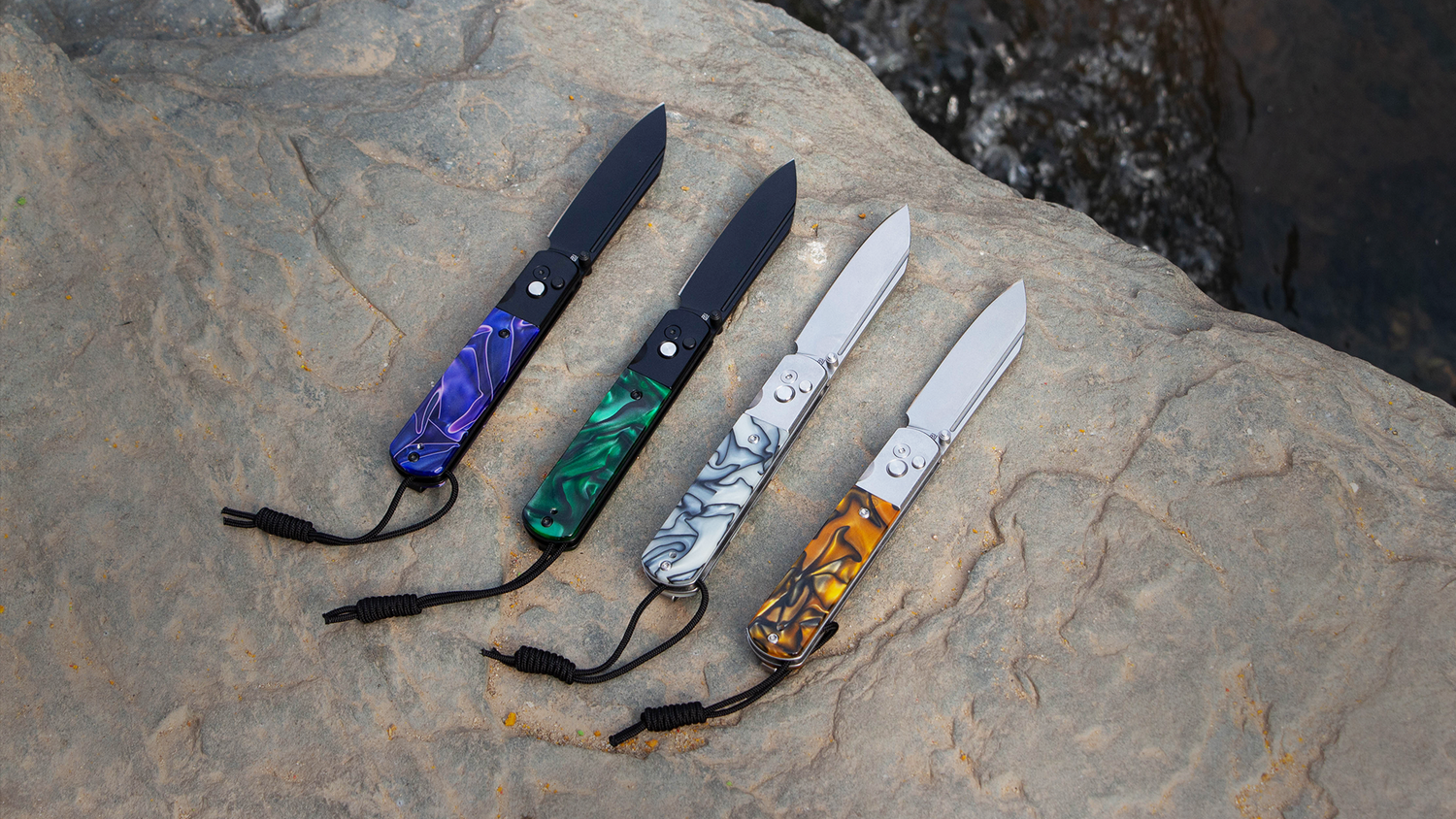 Four folding knives with colorful handles (blue, green, silver, and orange) arranged on a rock.